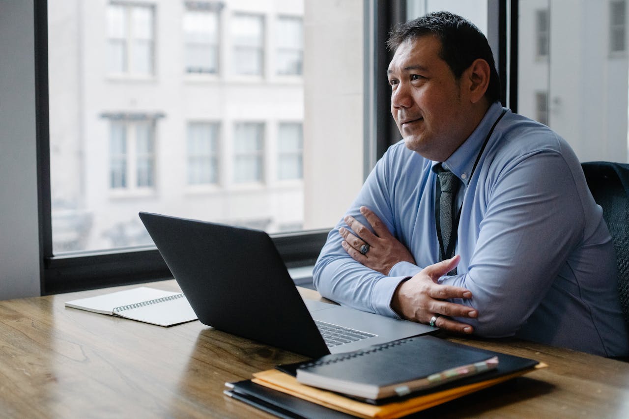 Adult successful ethnic male boss wearing shirt and tie sitting with hands crossed at workplace with documents and netbook